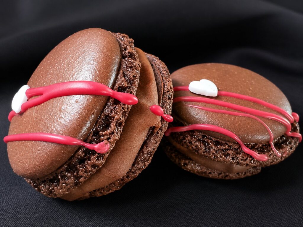 Two handcrafted chocolate French macarons decorated with pink drizzle and filled with rich chocolate ganache.