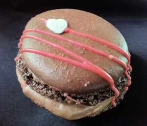 Handcrafted chocolate French macaron decorated with pink chocolate drizzle and a white heart, photographed on a black background.