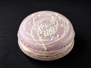 Handcrafted Earl Grey French macaron with delicate lavender-grey shells, creamy Earl Grey tea filling, white chocolate drizzle and crisp white pearls on a black background.