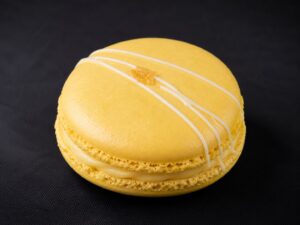 Handcrafted Lemon French macaron with bright yellow shells, creamy lemon filling, white chocolate drizzle and a touch of candied lemon on a black background.