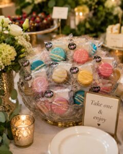 Elegant event display featuring individually wrapped Box of Nom macarons presented in a decorative glass bowl with flowers, candles and a "Take one & enjoy" sign for guests.
